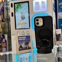 Photography of a black carbon fiber iPhone case with a magnetic ring holder and blue neon light accent, displayed next to its 'Vacuum adsorption Magnetic support Diamond Black' packaging box on a clear counter in a tech retail store