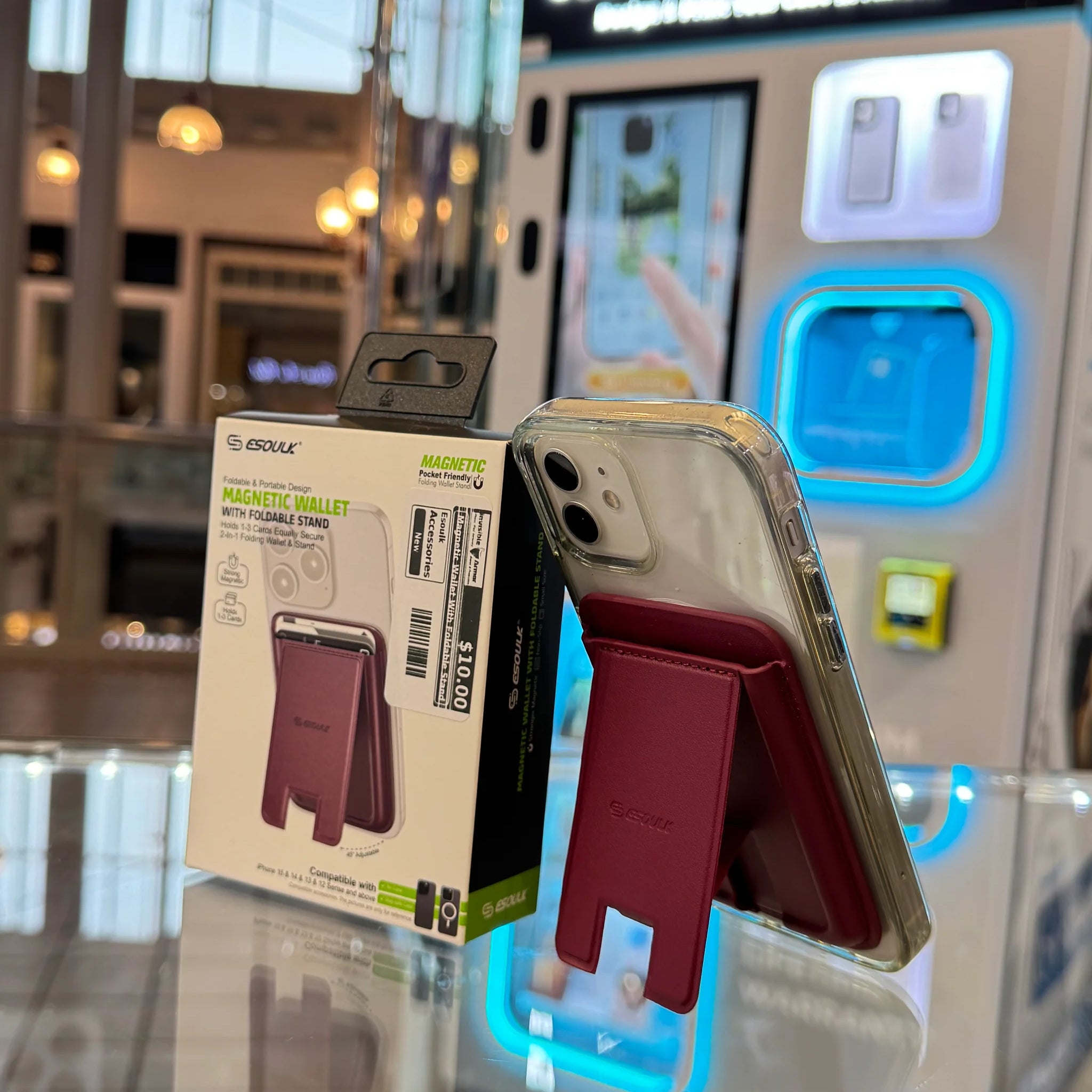Digital photograph of an ESSOULK magnetic maroon wallet phone stand attached to a clear-cased iPhone, displayed with its $10.00 packaging on a glass counter in front of a blue-glowing CUSTOM CASE KIOSK