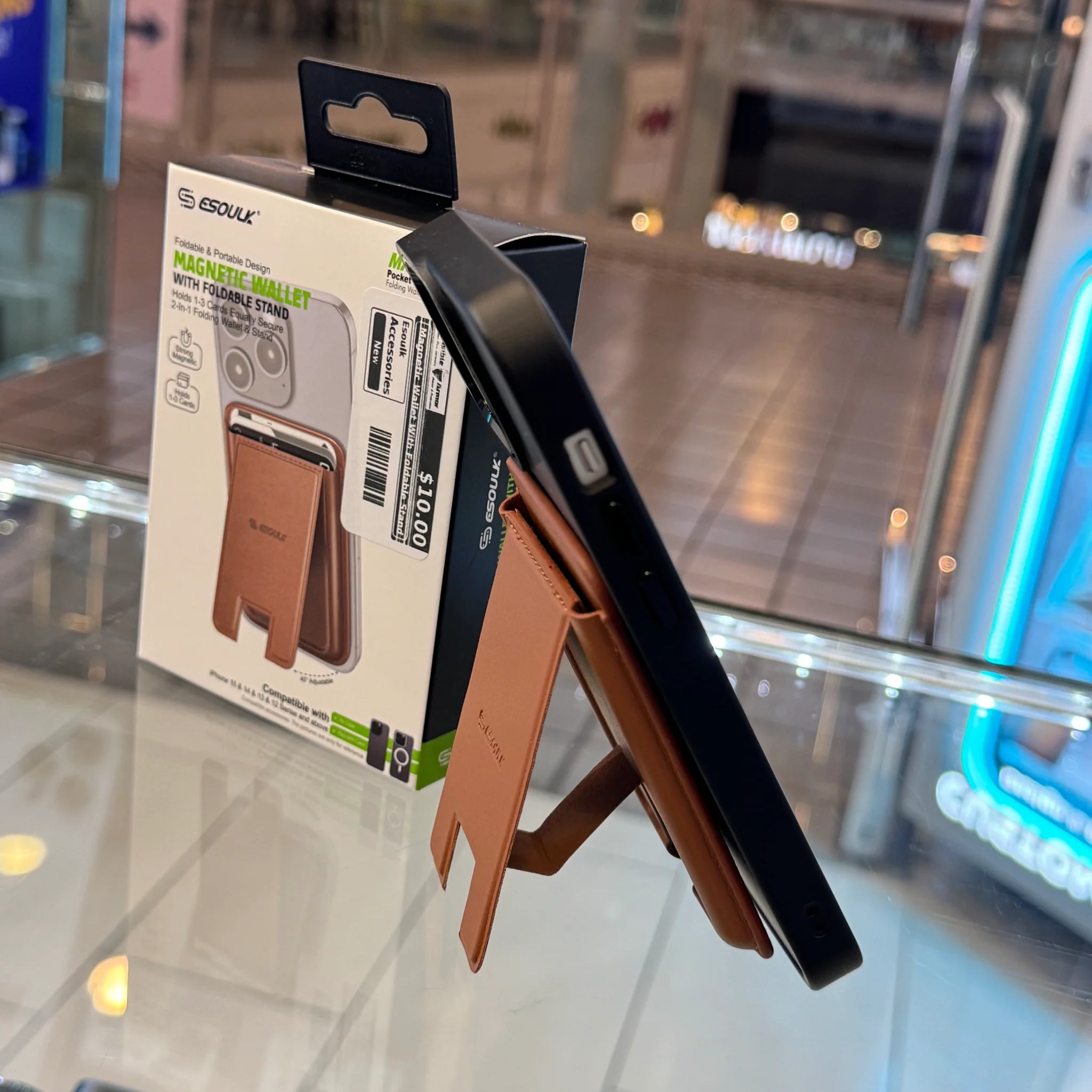 Photograph of an Esoulk brown leatherette magnetic wallet and foldable stand attached to a dark phone, displayed on a glass retail counter with its product packaging showing a $10 price sticker