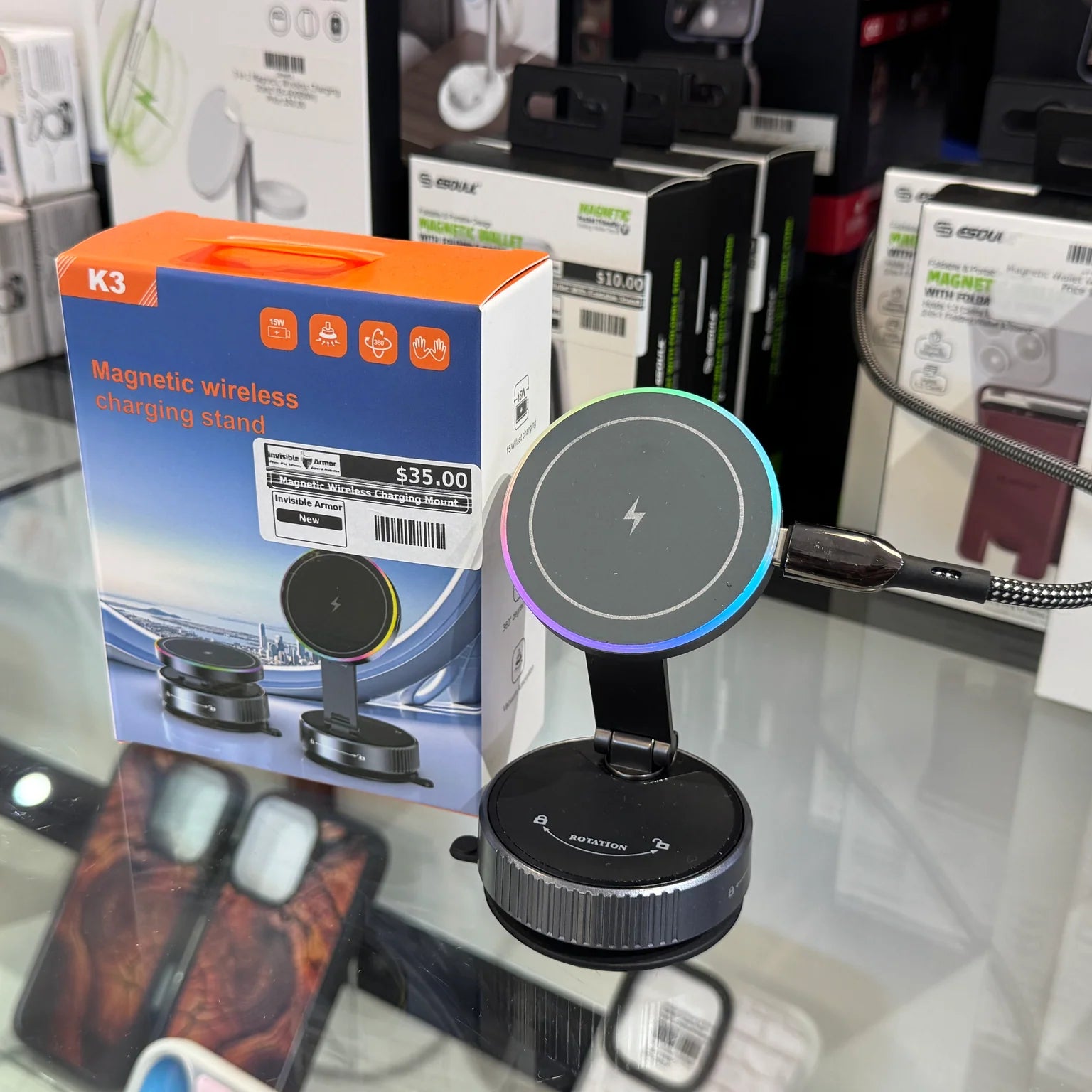 Photograph of a K3 Magnetic wireless charging stand featuring a colorful RGB light ring and a braided USB cable, displayed next to its orange and blue packaging with a $35.00 price tag on a glass retail counter
