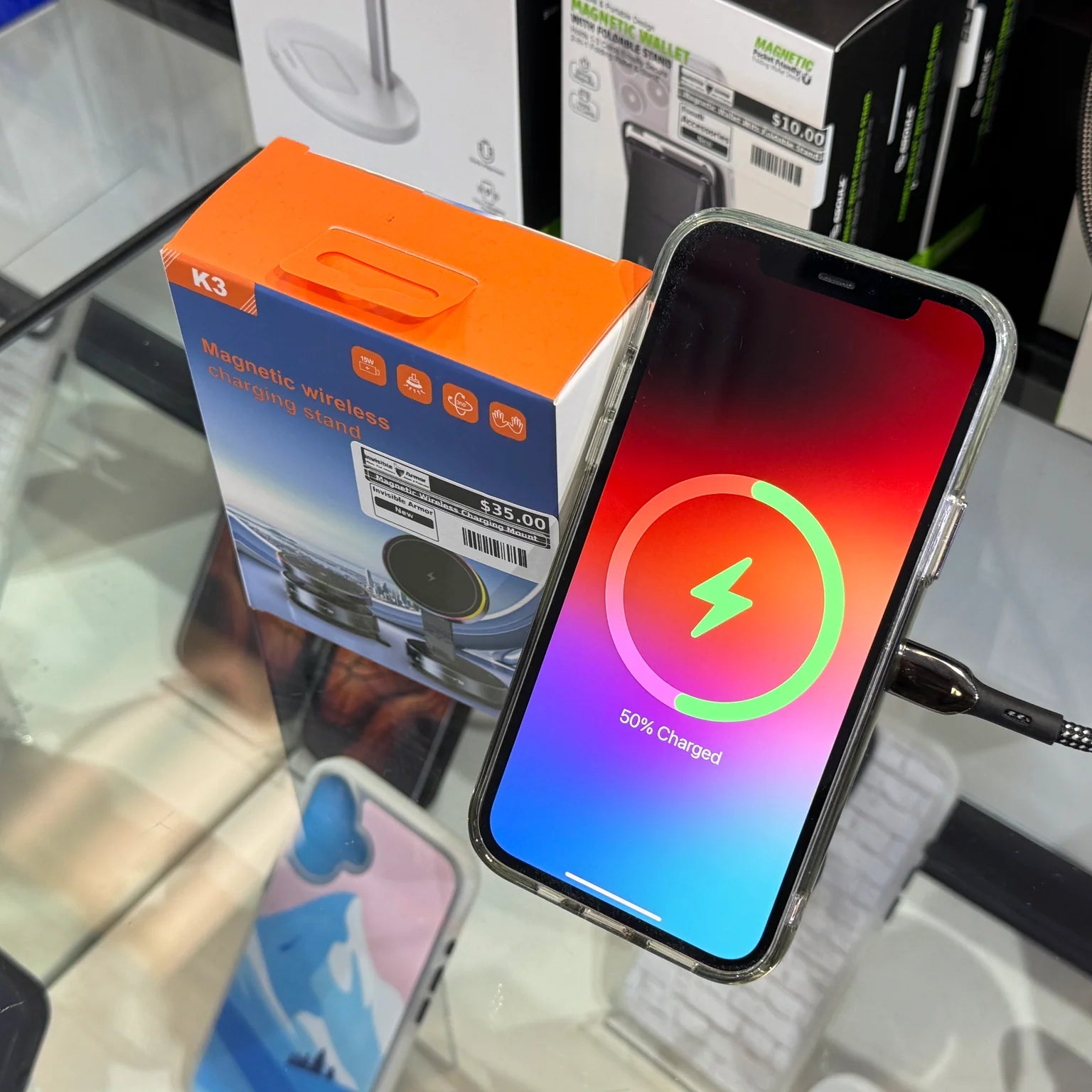 An iPhone displaying 50% charged with a vibrant red, pink, and blue gradient screen is plugged into a black braided cable, resting on a glass retail display next to an orange K3 Magnetic Wireless Charging Stand box priced at $35.00