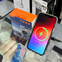 An iPhone displaying 50% charged with a vibrant red, pink, and blue gradient screen is plugged into a black braided cable, resting on a glass retail display next to an orange K3 Magnetic Wireless Charging Stand box priced at $35.00