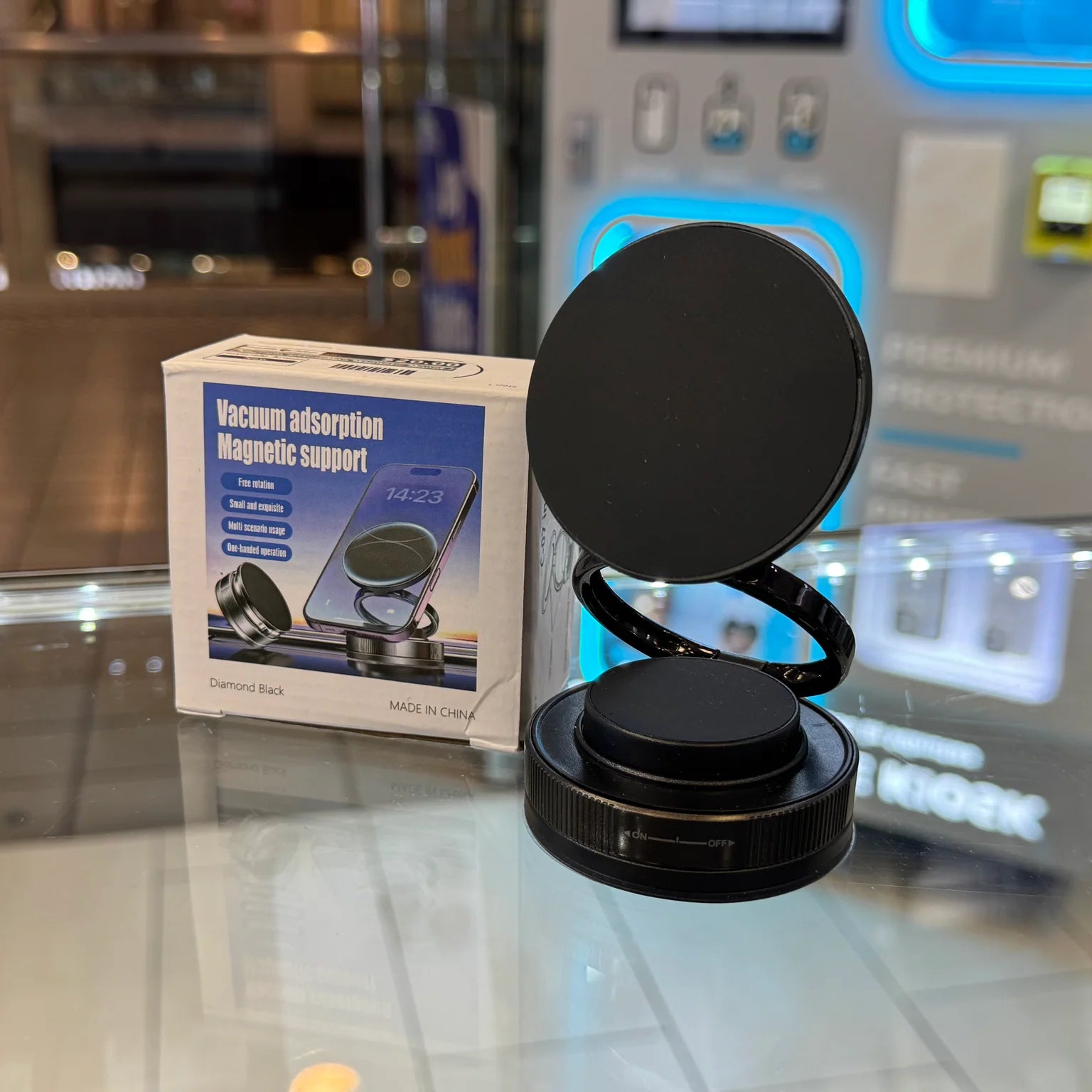 Photograph of a black magnetic phone support with a flexible coiled arm and its white packaging displaying a smartphone, resting on a glass counter in a retail store with blue neon lit displays