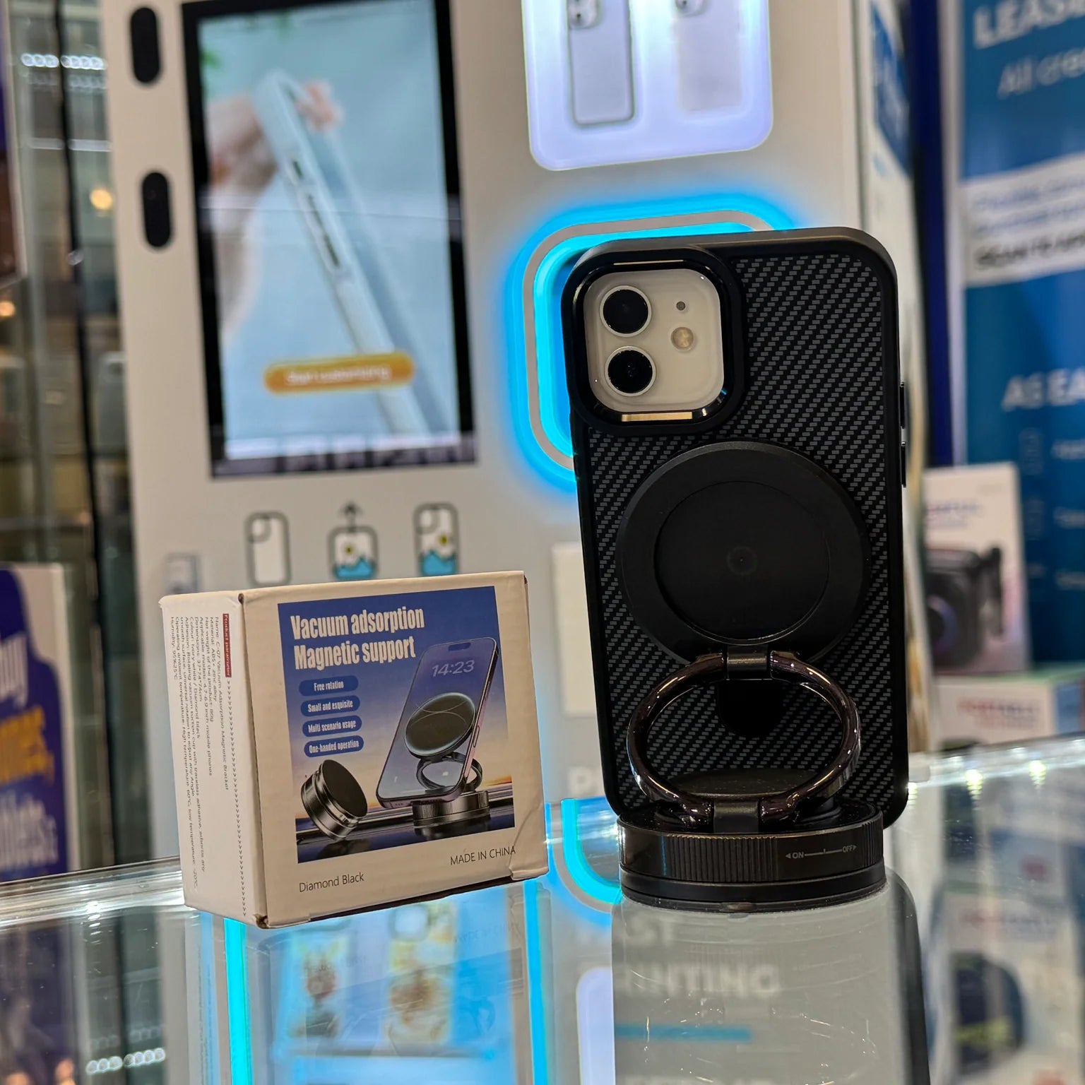 A black carbon fiber iPhone case with an integrated magnetic ring stand and its "Vacuum adsorption Magnetic support" product box sit on a glass counter in a retail electronics store with a blurred blue illuminated phone display in the background