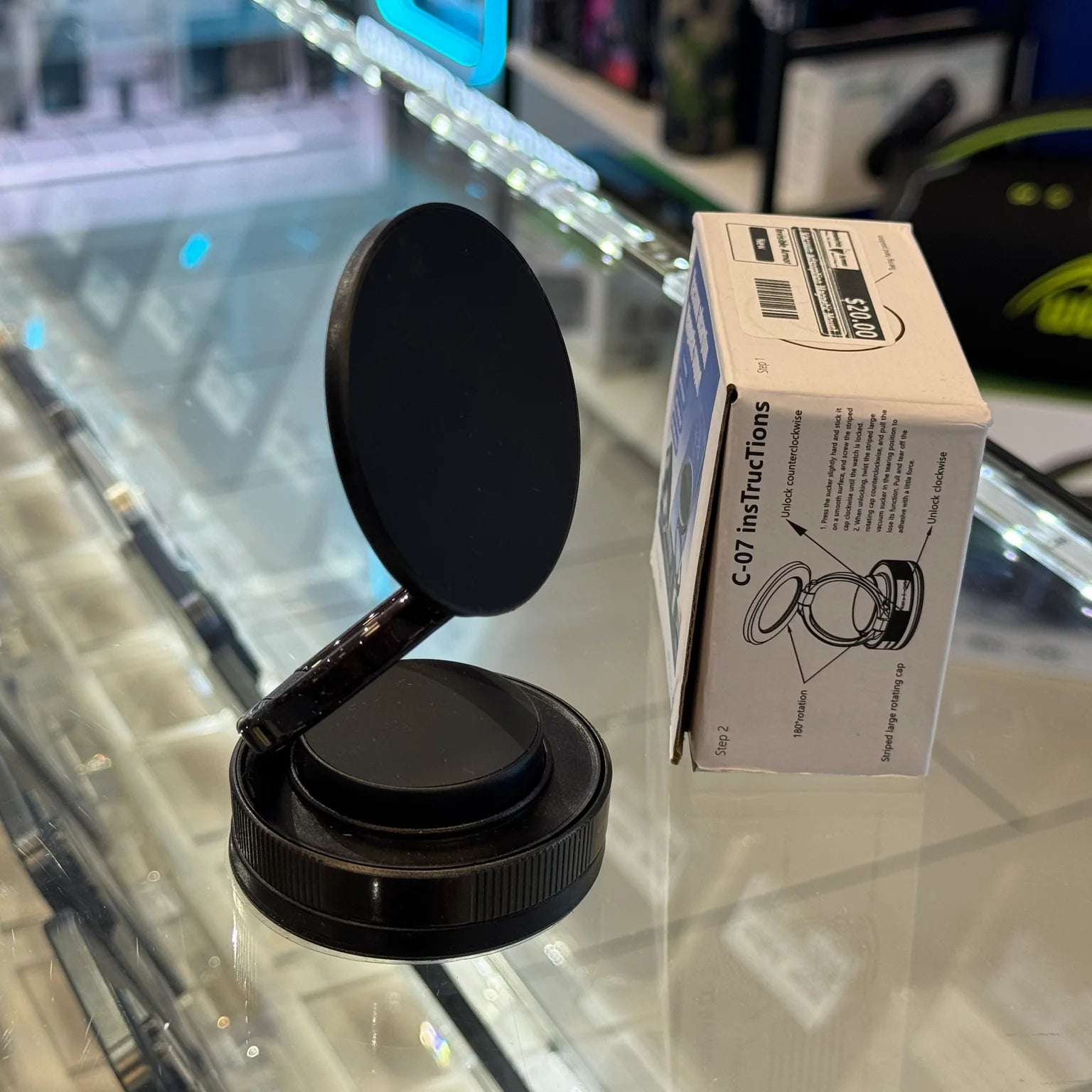 Photograph of a black magnetic phone mount next to its white C-07 instruction box featuring a $20.00 price sticker, displayed on a reflective glass counter with a blurred neon blue light and green logo in the background