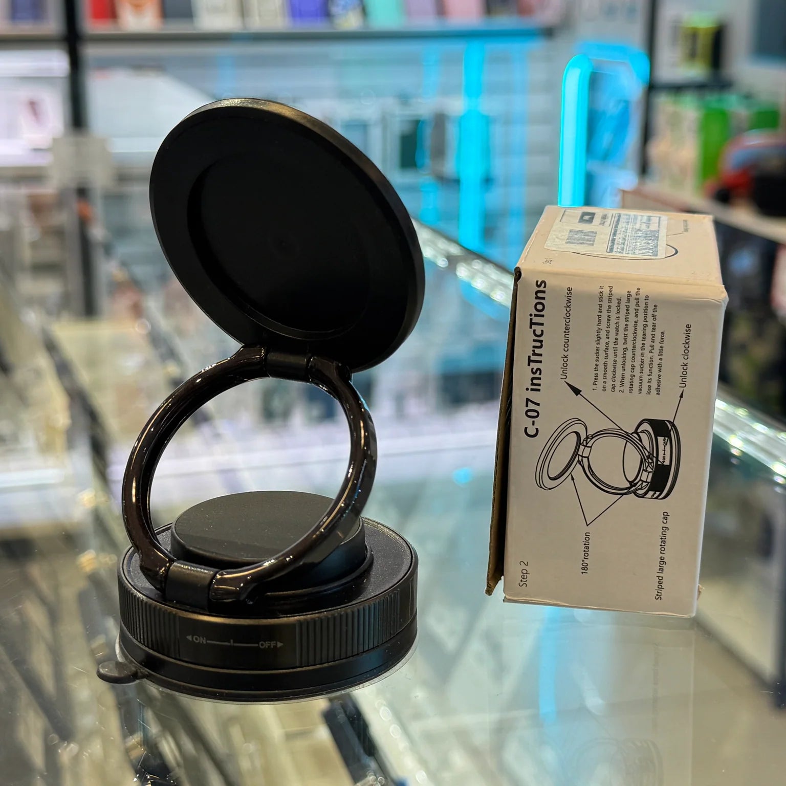 Photograph of a sleek black phone holder with a rotating ring and suction cup base resting on a reflective glass counter in an electronics store, next to a white instruction box detailing its C-07 model features, with a blurry vibrant blue neon light in the background