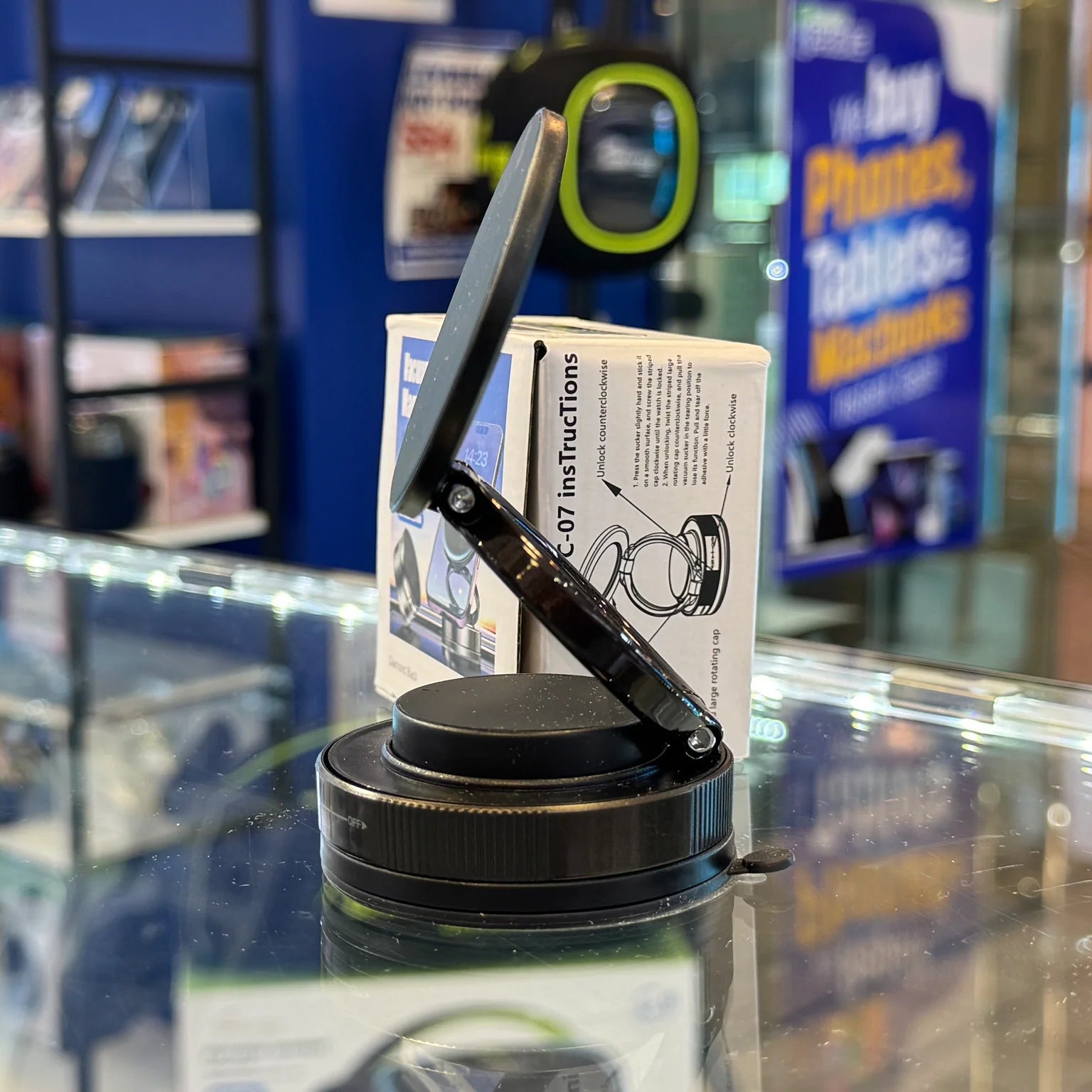 Black adjustable phone stand angled on a reflective glass display counter in an electronics store, with a white instruction box and blurry blue signs for phones and tablets behind it