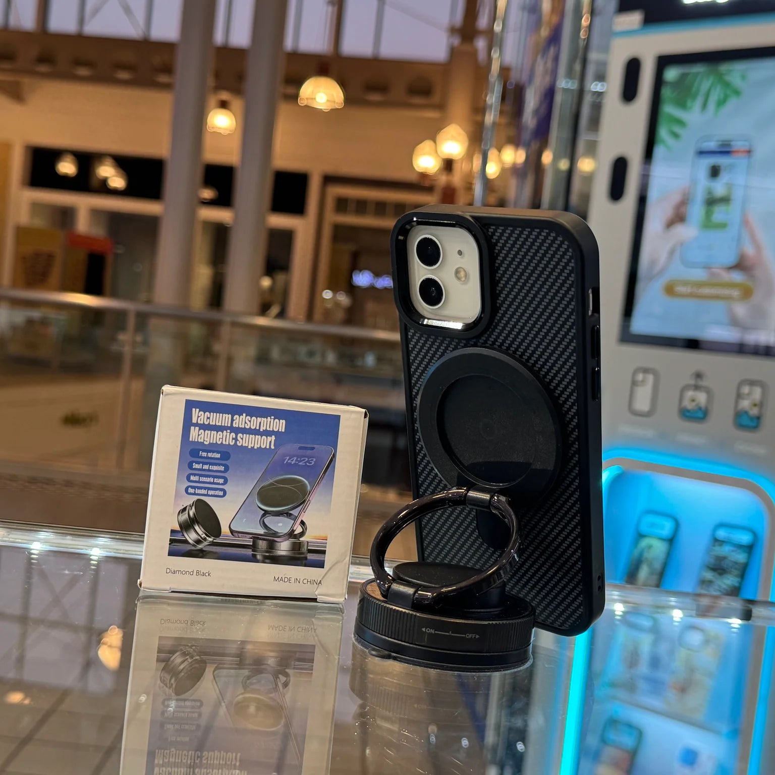 A black iPhone in a textured case with a magnetic ring holder sits on a reflective surface alongside its product box for Vacuum adsorption Magnetic support Diamond Black inside a brightly lit retail environment featuring glowing blue display units and a blurred reflected JCPenney sign
