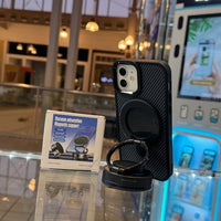 A black iPhone in a textured case with a magnetic ring holder sits on a reflective surface alongside its product box for Vacuum adsorption Magnetic support Diamond Black inside a brightly lit retail environment featuring glowing blue display units and a blurred reflected JCPenney sign