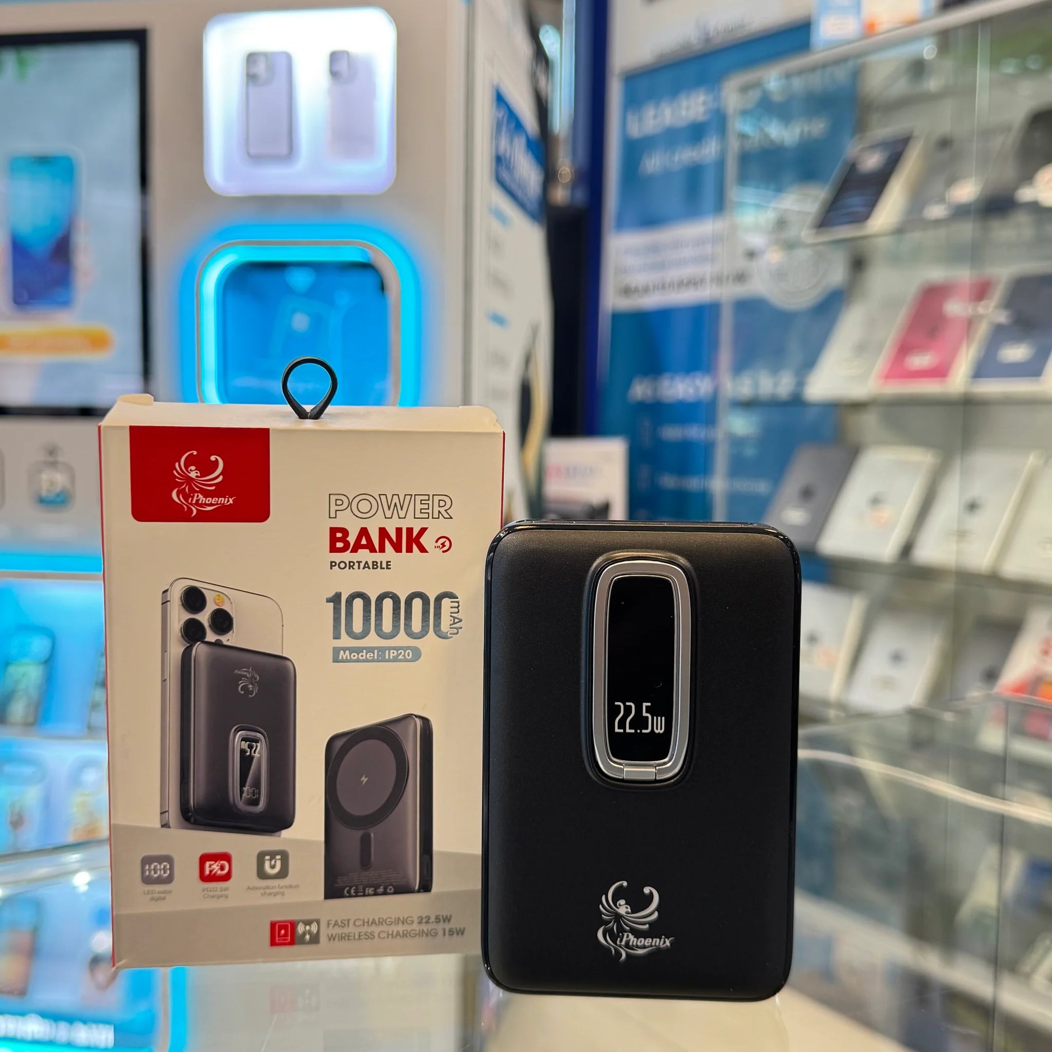 A black iPhoenix portable power bank with a silver-framed 22.5W digital display rests next to its white 10000mAh packaging on a reflective counter in a retail electronics store featuring a glowing blue CUSTOM CASE KIOSK and blurred shelves of phone accessories in the background