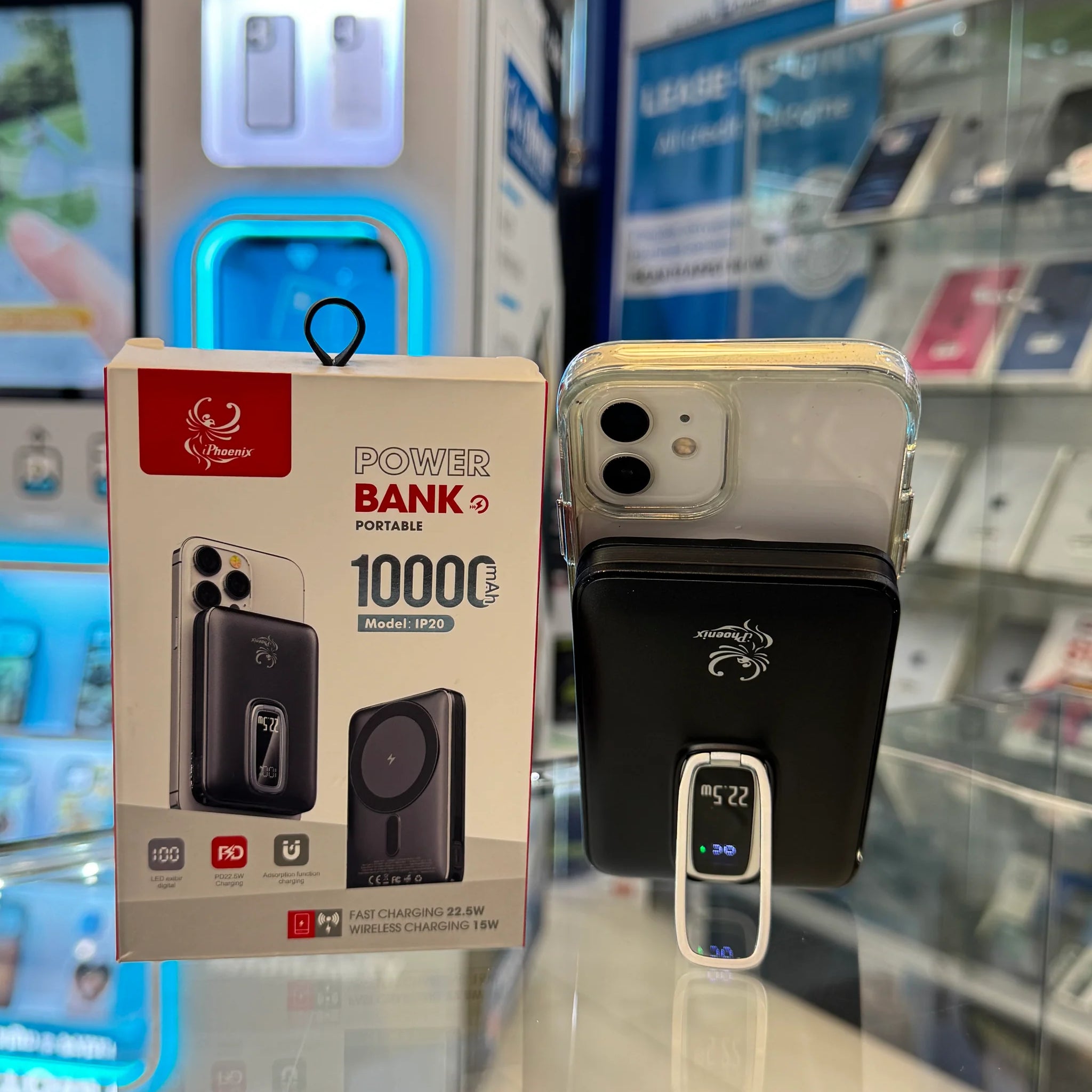 Digital photograph showing a Phoenix 10000mAh portable power bank, presented in its box with a red logo and also attached to a white iPhone displaying 22.5W, all within a brightly lit Custom Case Kiosk with blue display accents