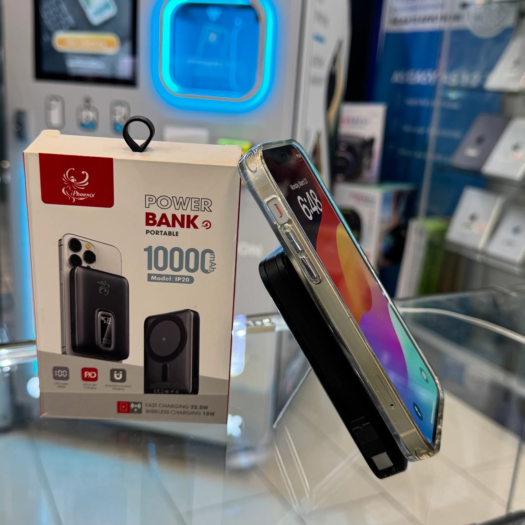 Retail display featuring an iPhoenix 10000 mAh portable power bank box with a red phoenix logo and an iPhone in a clear case magnetically attached to a black charger, all on a glass shelf against a background of blue illuminated display cabinets