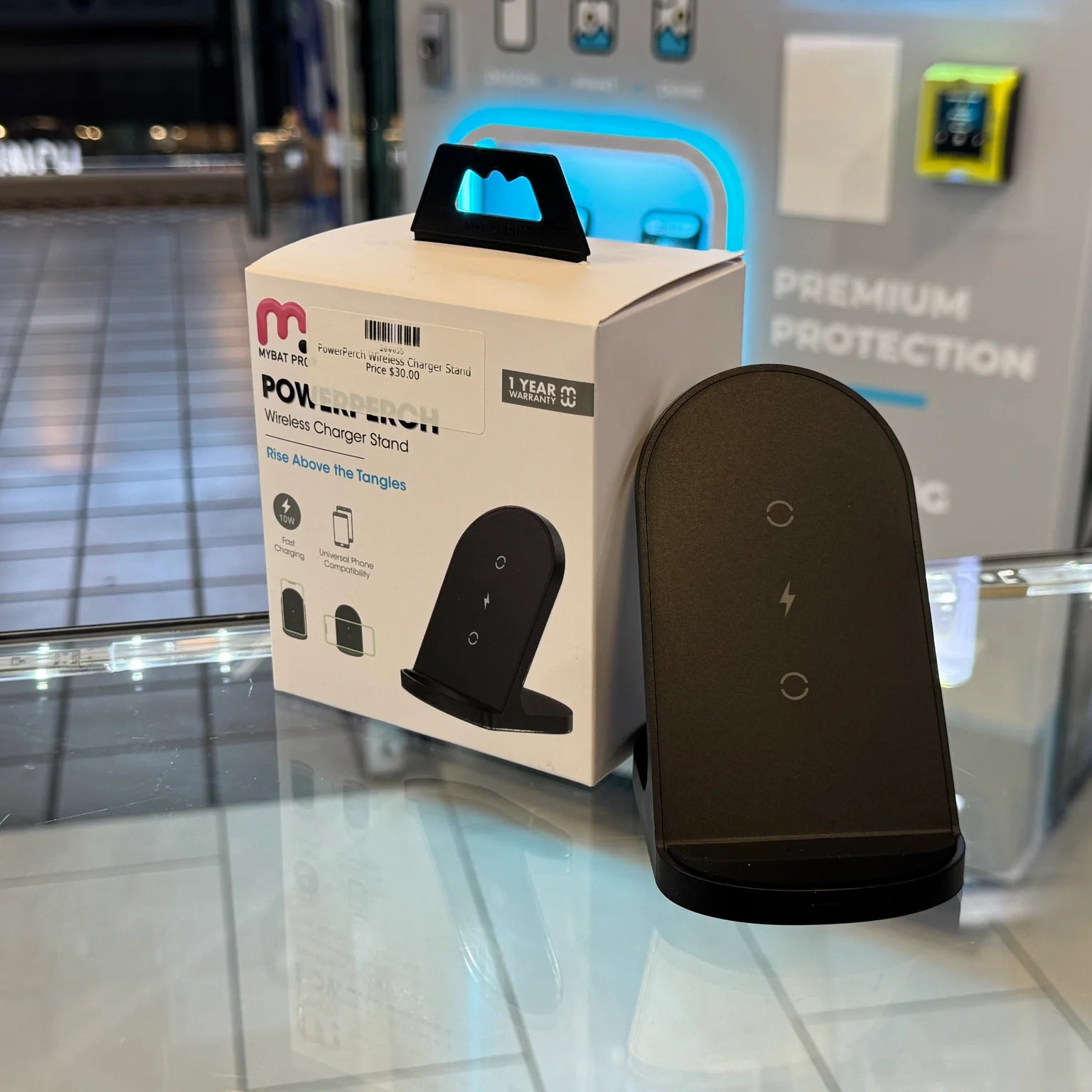 A MYBAT PRO PowerPerch wireless charger stand on a glass retail display, showing its white product box with a $30.00 price tag and "1 YEAR WARRANTY" badge, next to the black charger device, against a backdrop of a glowing blue light and "PREMIUM PROTECTION" text