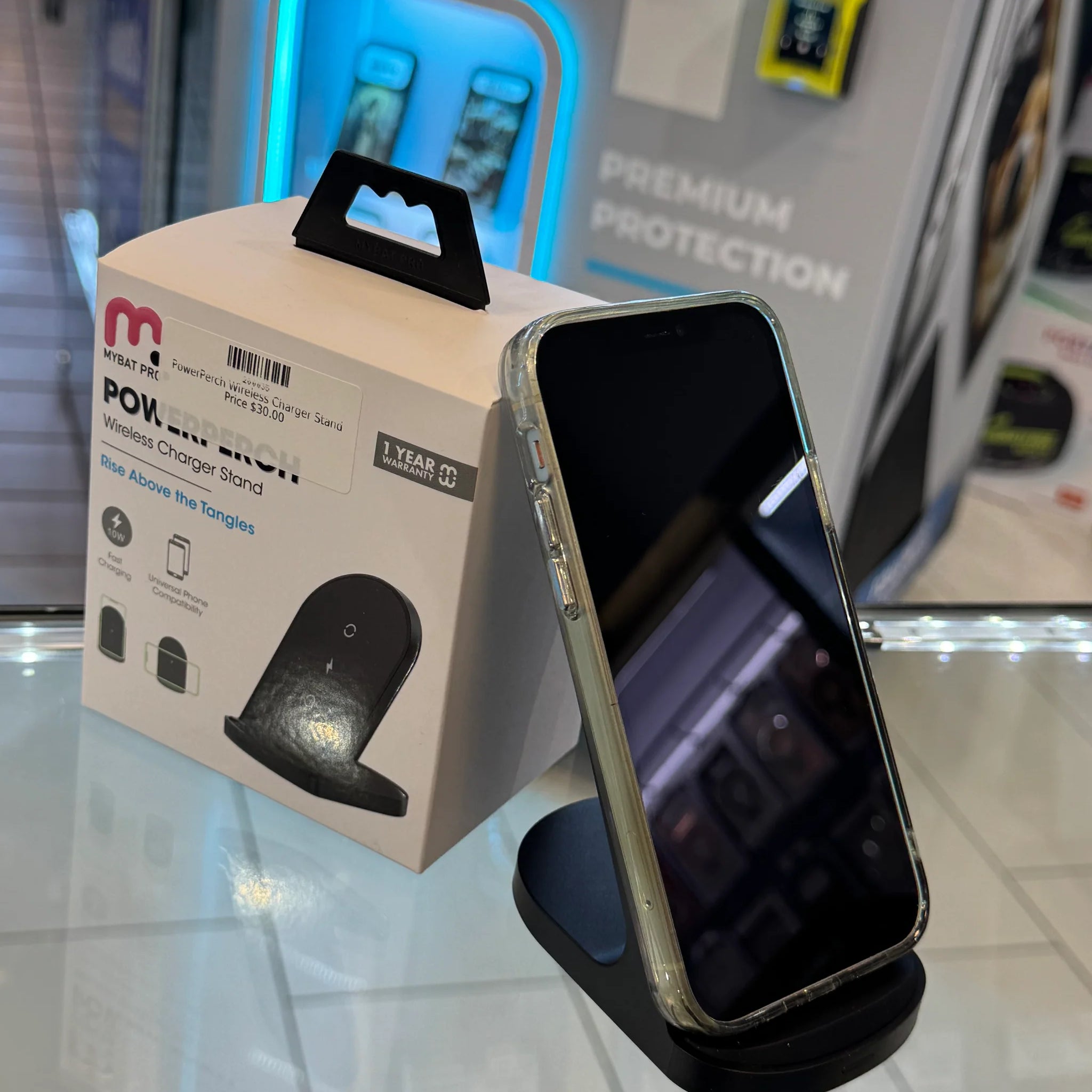 A black MyBat Pro PowerPerch wireless charger stand holding an iPhone in a clear case, showcased on a retail display with vibrant blue LED lighting and a "Premium Protection" sign