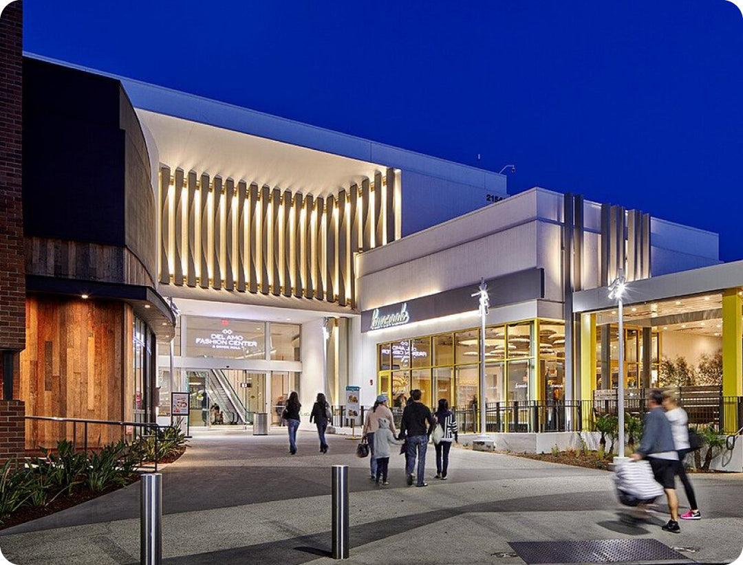 Modern shopping mall exterior with illuminated storefronts and people walking outside.